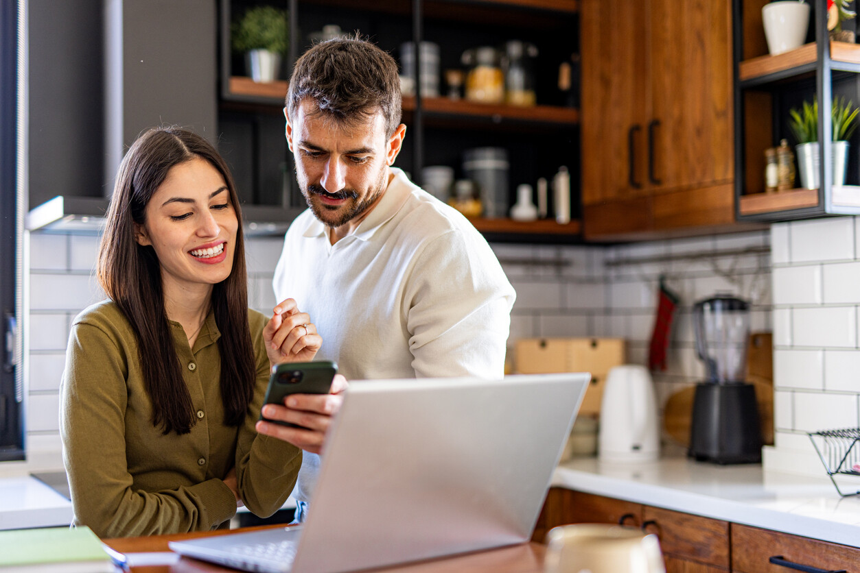 Young smiling couple using laptop and smartphone, checking their home finances in the kitchen